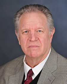 "portrait of a man in a suit and tie set against a muted background. He's shown from shoulders up"