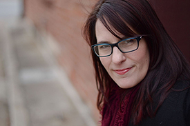 "portrait of a woman shown from shoulders up. she's set against a brick background. Wears plastic rimmed glasses and medium length brown-ish red-ish hair"