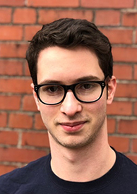 young man in plastic, black glasses with dark short hair looks directly at the camera smiling slightly. Behind him is a brick wall