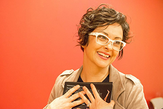 "portrait of a woman clutching a book to her chest and looking off camera. She wears glasses and a smile."