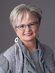 Woman dressed in gray with wire-rimmed glasses. She smiles widely and has short gray hair.